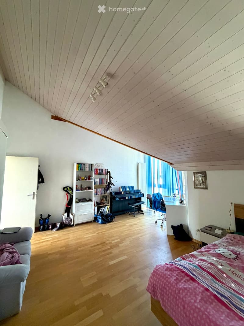 Bright attic bedroom with wooden floors, slanted wooden ceiling, large windows, bed with pink bedding, sofa, bookshelf, piano, and desk area.