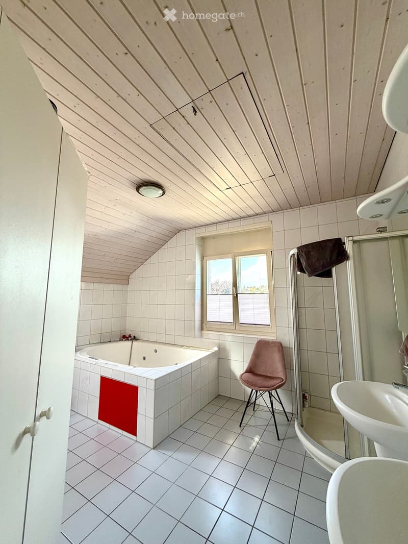 Bathroom with white tiled walls and floor, a bathtub with a red towel, a sink, a pink stool, and a shower area with a towel hanging on the side.