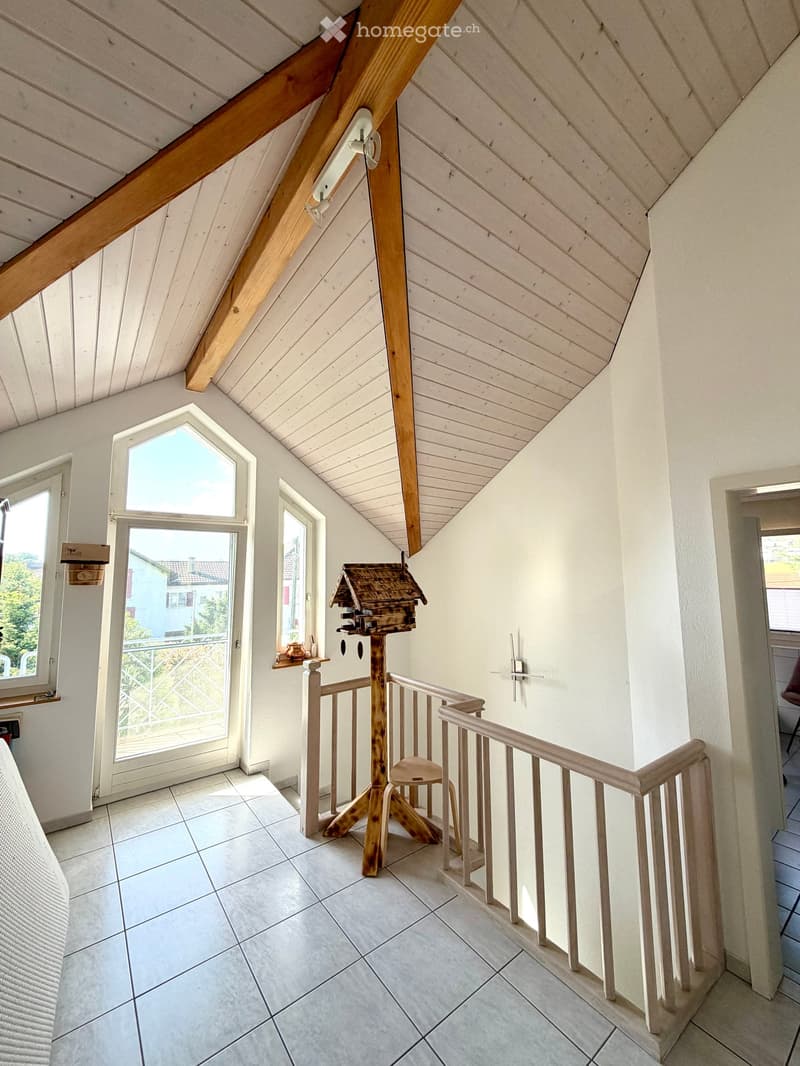 Bright hallway with white walls, wooden ceiling beams, and a staircase leading upwards. Features include a bird feeder on a stand, a small stool, and windows offering a view of the outdoors.