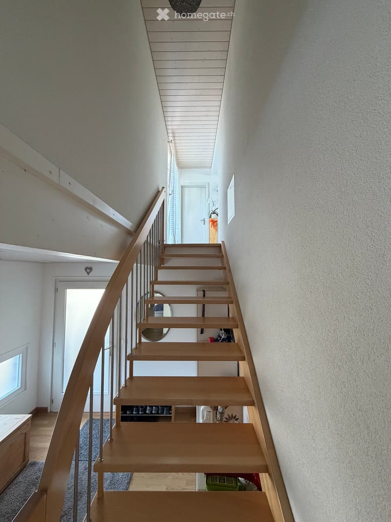 Modern wooden staircase with metal railing, leading to an upper level. The walls are painted in a light neutral tone, and there is a small window on the left side providing natural light. A shoe rack is visible at the bottom of the stairs, and a door is s