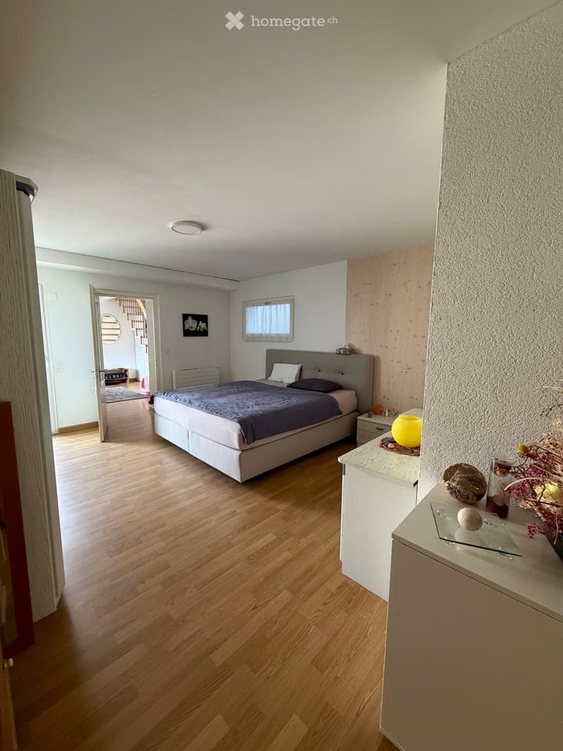 Modern bedroom with wooden flooring, a large bed with dark bedding, a nightstand with a lamp, and a small table with decorative items.