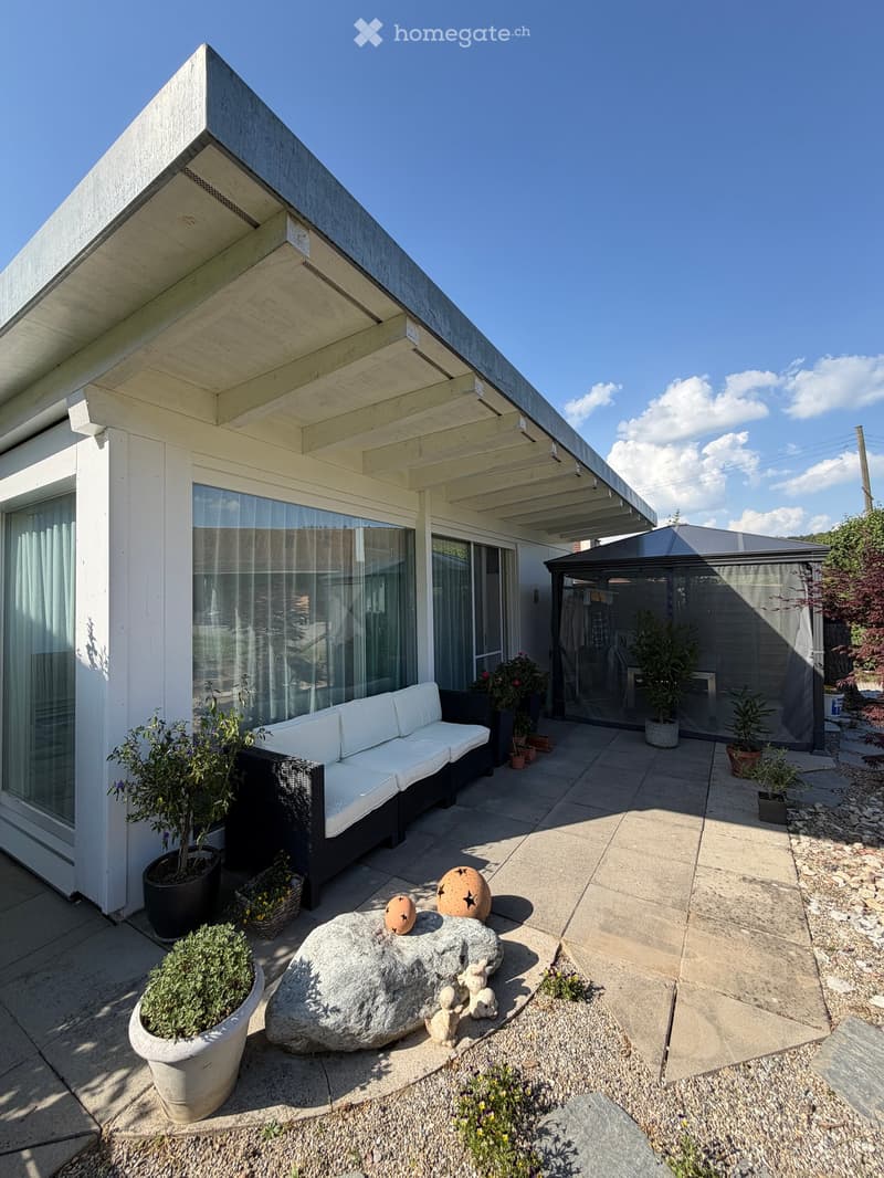 Modern outdoor patio with a white sofa, decorative rock, potted plants, and a covered seating area with glass walls.