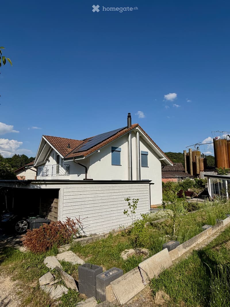 Two-story house with a tiled roof, solar panels, and a garage. The house has white walls, blue shutters, and a small balcony on the upper level.