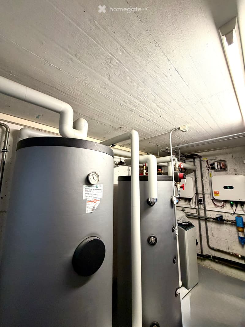 Utility room with two water heaters, pipes, valves, and a control panel mounted on the wall.