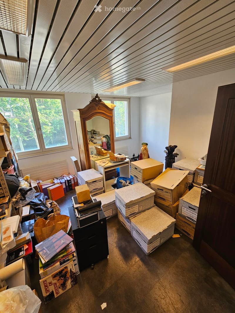An attic room with slanted ceilings, large windows, and various items including a mirror, desk, chair, and stacked cardboard boxes.