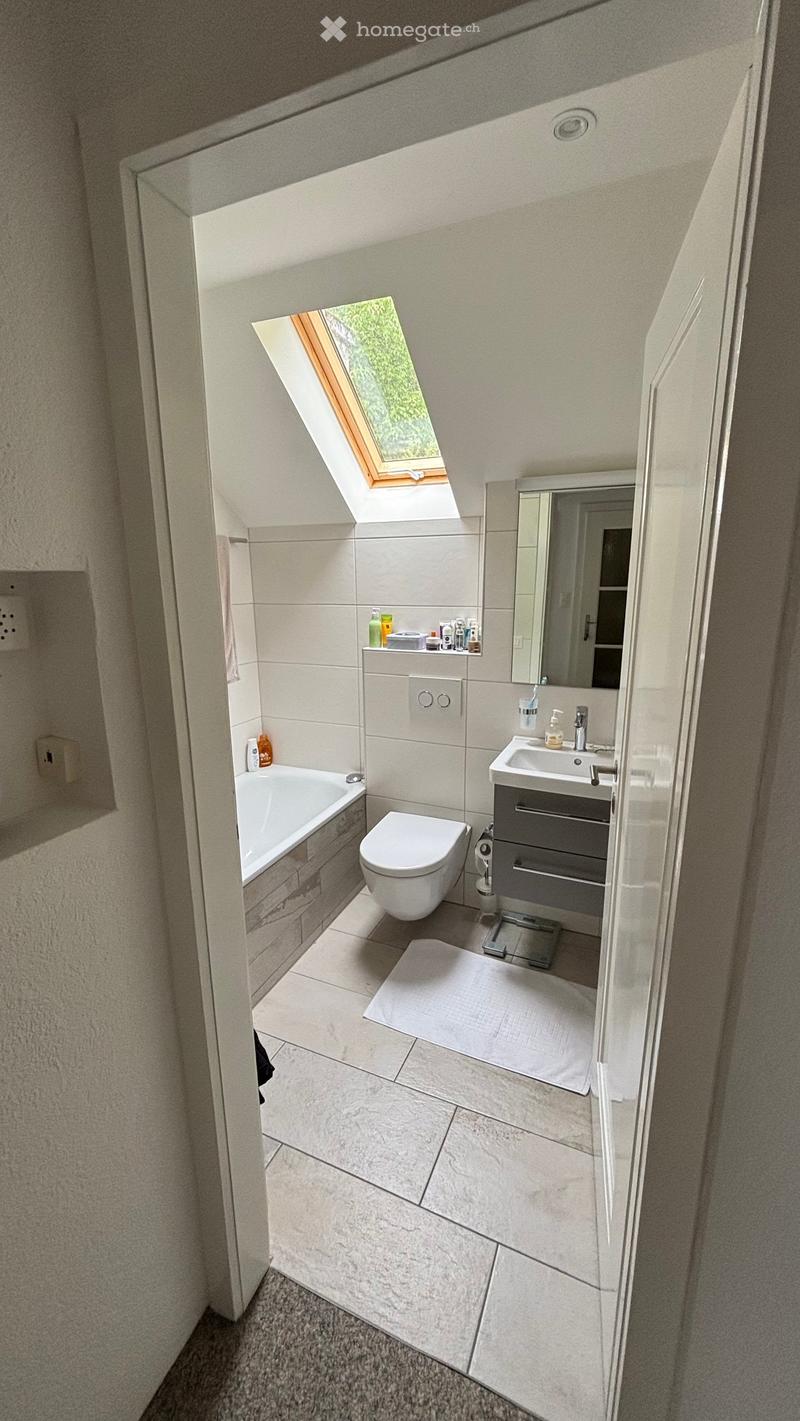 Modern bathroom with a bathtub, toilet, sink, mirror, and cabinet. Features a skylight window, tiled walls, and a small white rug on the floor.