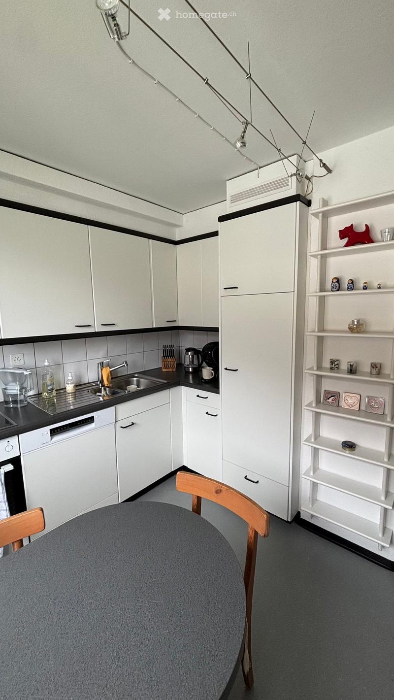Modern kitchen with white cabinets, black countertops, and stainless steel appliances. Includes a round table with wooden chairs, a shelf with decorative items, and various kitchen utensils.