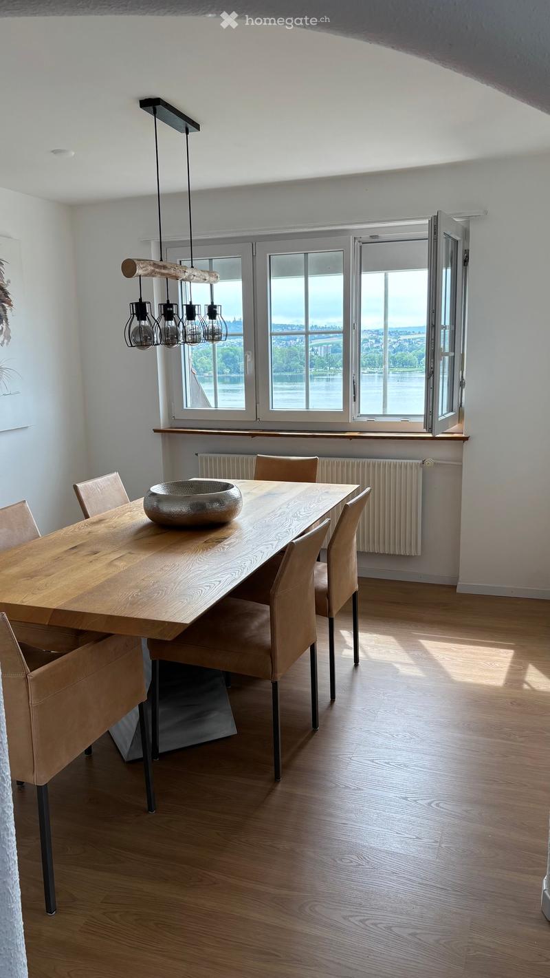 Bright dining area with wooden floor, large windows offering a view, a wooden dining table with six chairs, a decorative bowl, and a modern pendant light fixture.