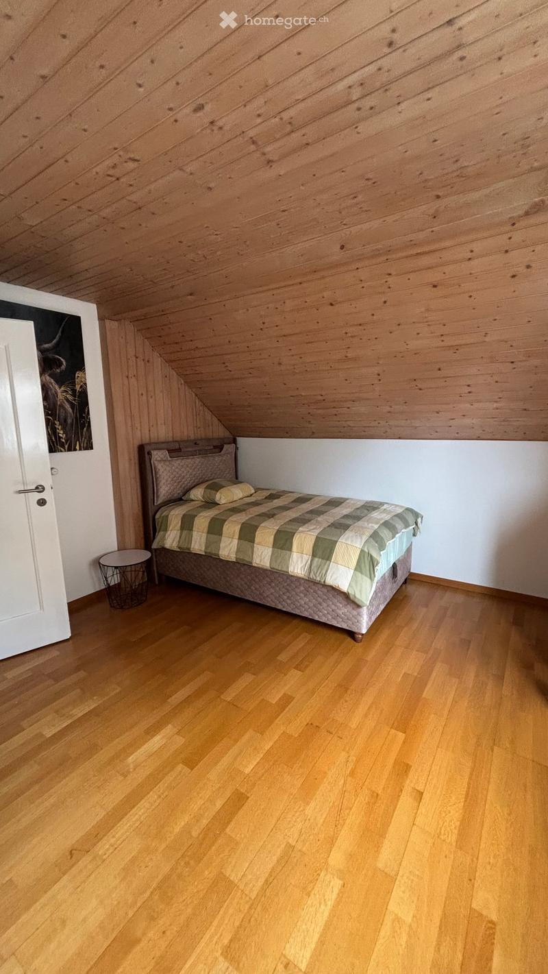 Cozy attic bedroom with wooden slanted ceilings, hardwood floors, a neatly made bed with a plaid duvet, a small round nightstand, and a framed picture on the wall.