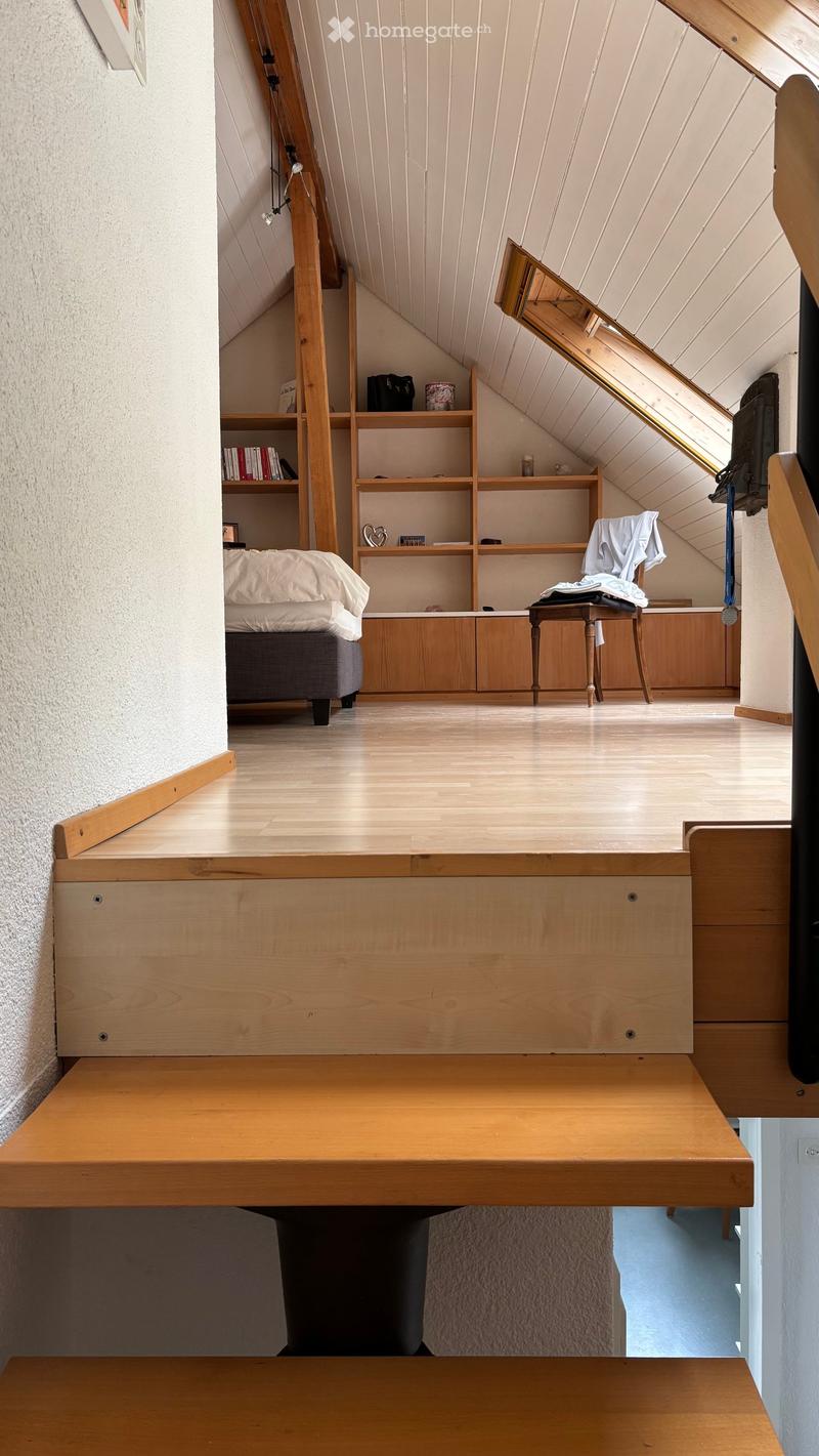 Cozy attic bedroom with wooden floors, slanted ceilings, built-in wooden shelves, a small bed, a wooden stool, and a small table.