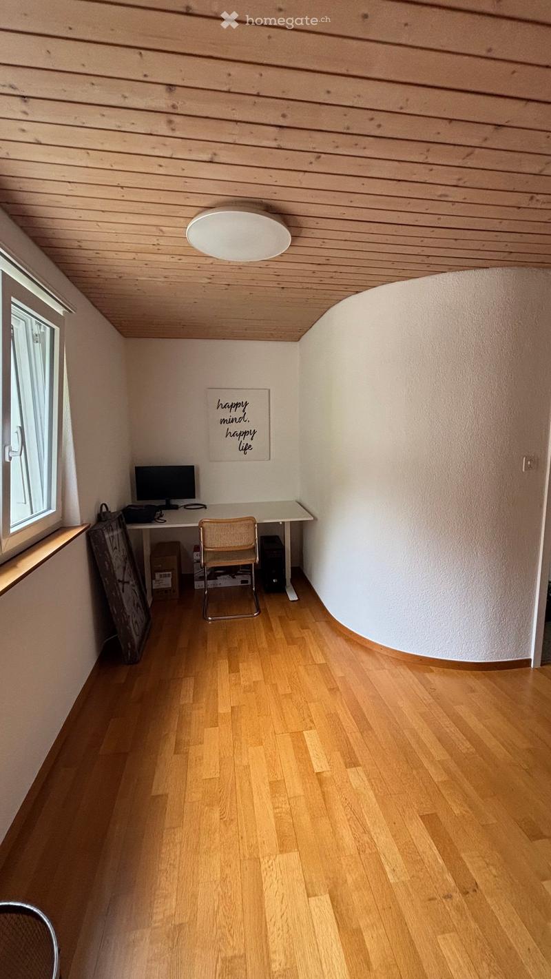 A small, well-lit room with wooden flooring and a slanted wooden ceiling. Features a desk with a computer monitor, a chair, and a decorative sign on the wall.