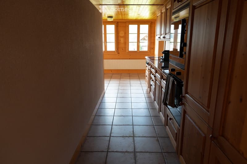 Bright kitchen with wooden cabinets, tiled floor, and large windows letting in natural light. Features an oven, coffee maker, and a towel hanging on the oven handle.