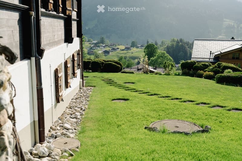 A well-maintained garden with a stone border, green grass, and a view of a village and mountains in the background.