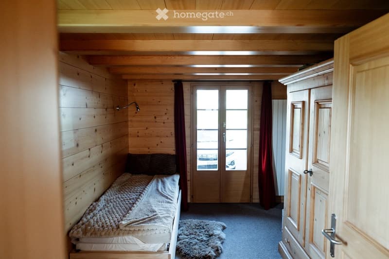 Cozy wooden bedroom with a single bed, wardrobe, and a furry rug on the floor. The room features wooden walls and a large window with curtains.