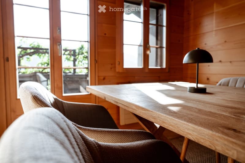 A cozy, wooden-furnished room with a large wooden table, a modern lamp, and a comfortable chair, featuring large windows with wooden frames that let in natural light.