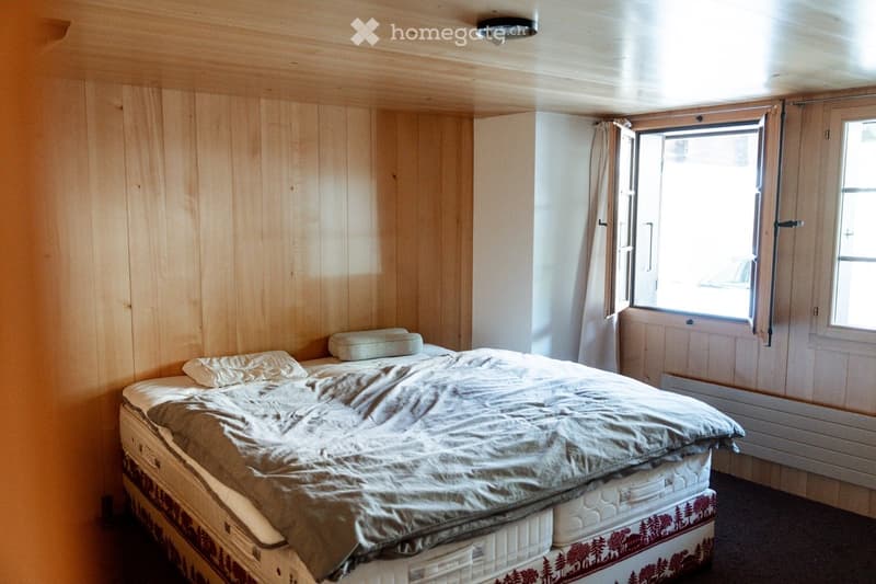 Cozy bedroom with wooden walls and ceiling, featuring a neatly made bed with pillows and a duvet, and a large window with curtains allowing natural light to enter.