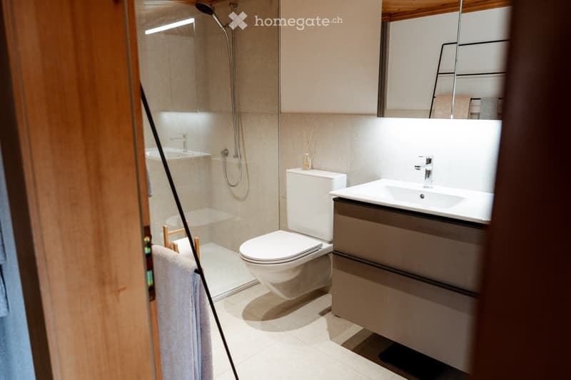 Modern bathroom with a white toilet, sink, and glass-enclosed shower. Features a large mirror, wooden accents, and a towel hanging on the door.