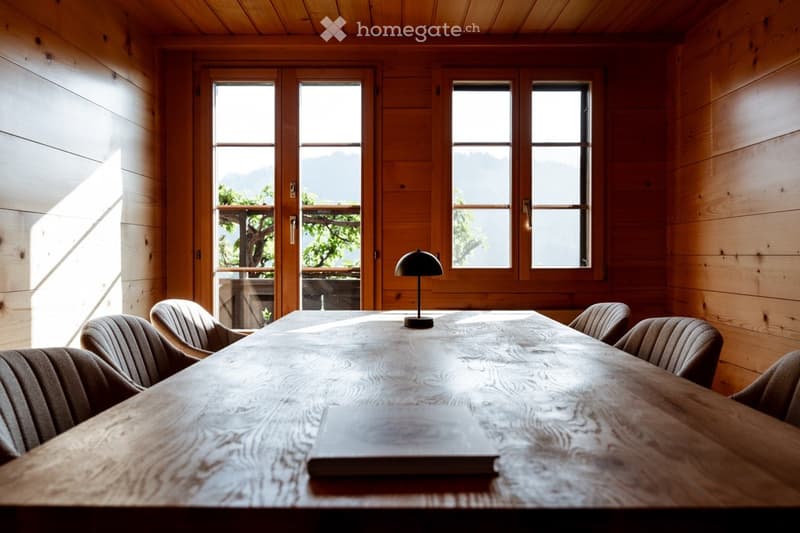 Bright, wooden-paneled room with large windows offering a scenic mountain view. A long wooden table is surrounded by several chairs, and a sleek black lamp sits on the table.