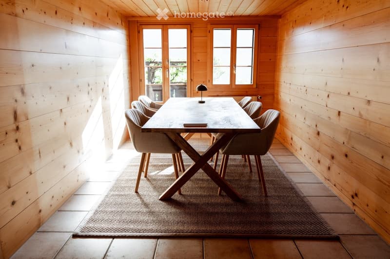 Wooden dining room with a large wooden dining table, six chairs, and a small table lamp on the table. The room features wooden walls and a wooden floor, with large windows and a sliding glass door leading to an outdoor area.