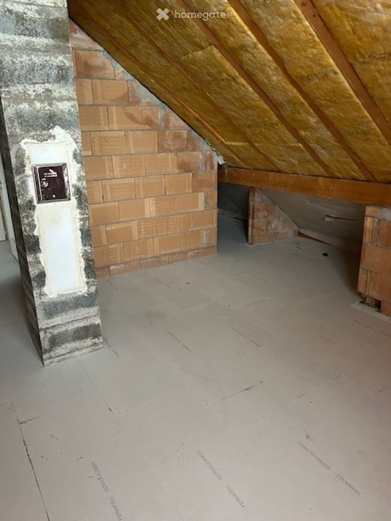 Empty attic space with exposed wooden beams, brick wall, and concrete floor. No furniture or equipment present.