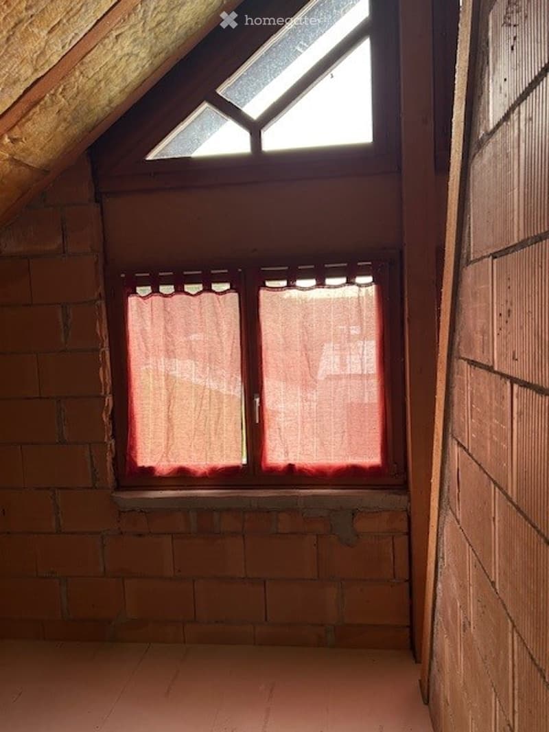 Empty attic room with exposed wooden beams, brick walls, and a skylight window with red curtains.