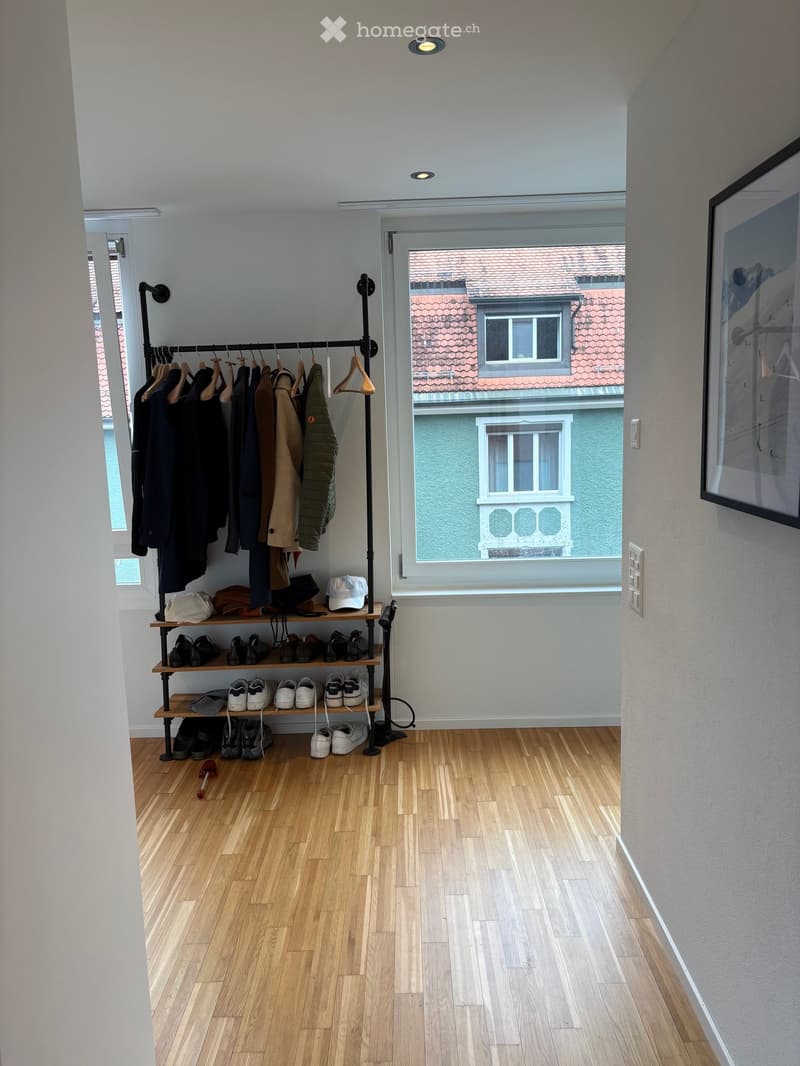 Empty hallway with wooden flooring, coat rack with jackets and coats, shoe shelf with multiple pairs of shoes, and a large window showing a view of neighboring buildings.