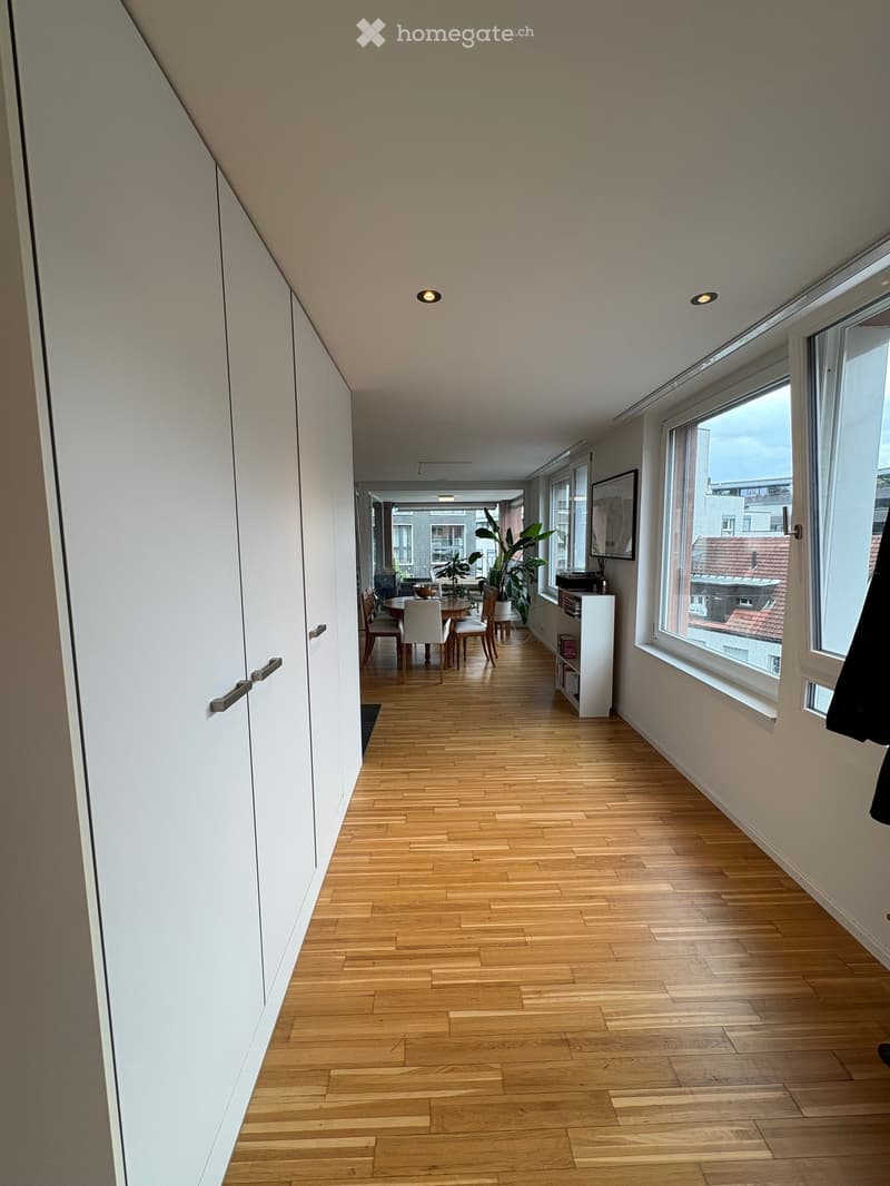 Bright hallway with wooden flooring, white walls, large windows, and recessed lighting. A small dining area is visible at the end with a table and chairs.