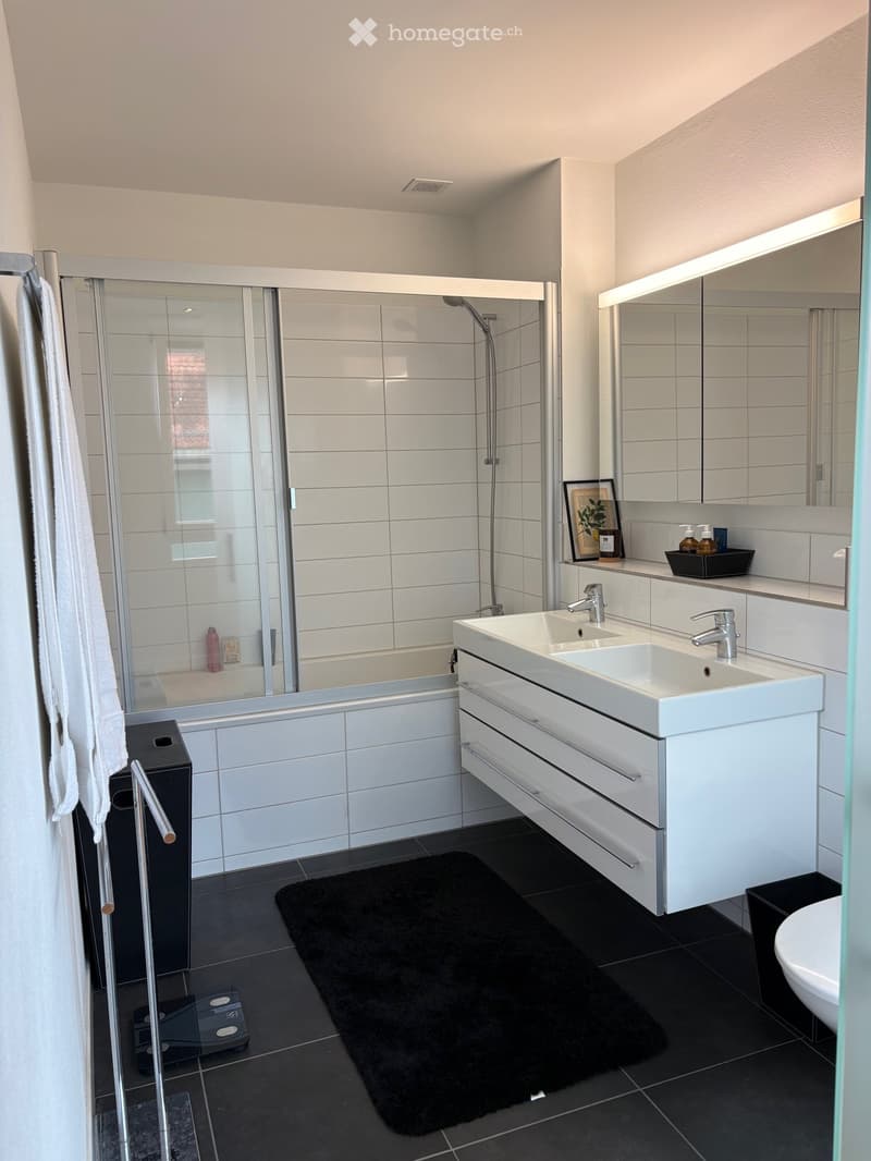 Modern bathroom with white tiles, a bathtub, a double sink, a large mirror, and a black bath mat.