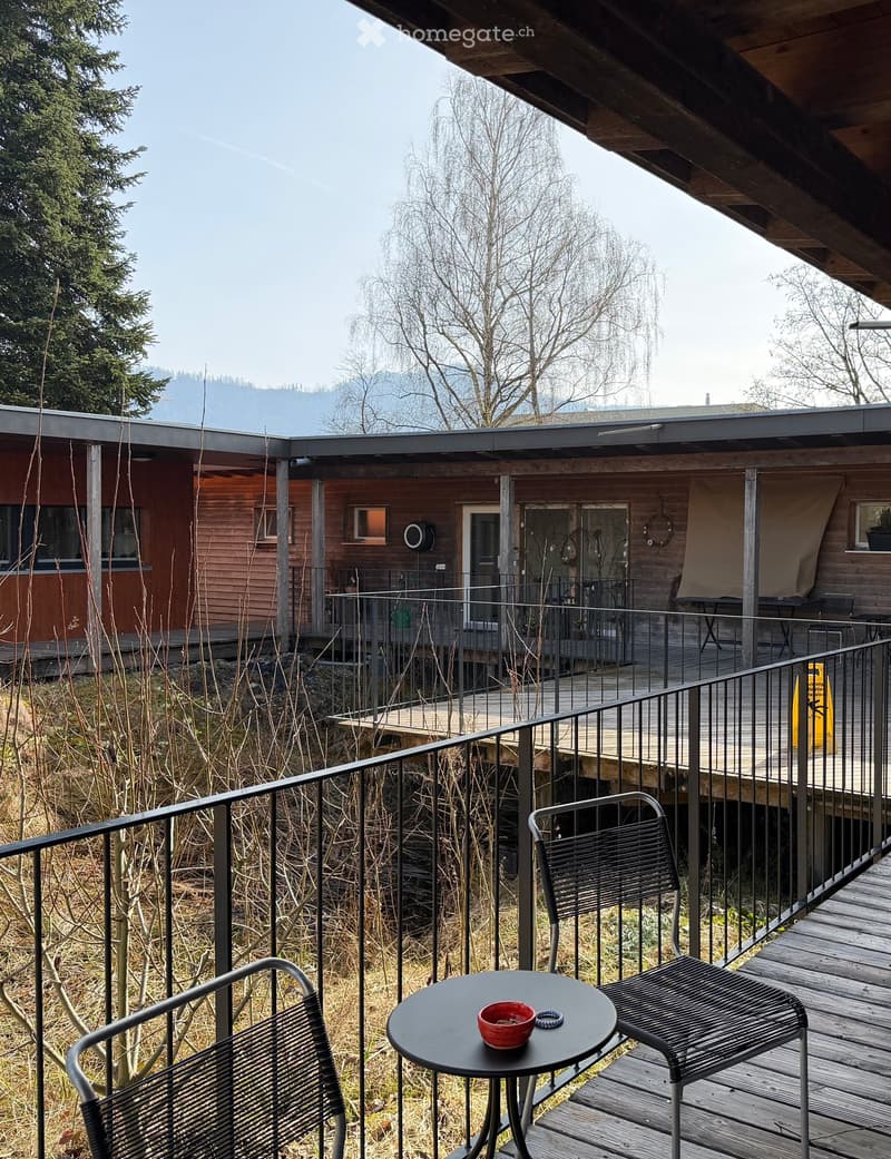 Wooden balcony with metal railings, two chairs, a small table, and a bowl on it.