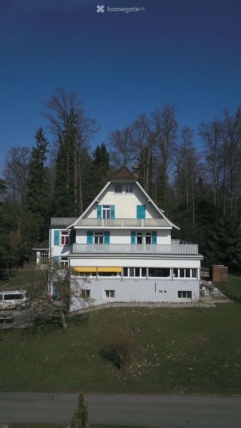 Two-story house with white walls, blue shutters, and a brown roof. It is surrounded by a grassy area and trees.