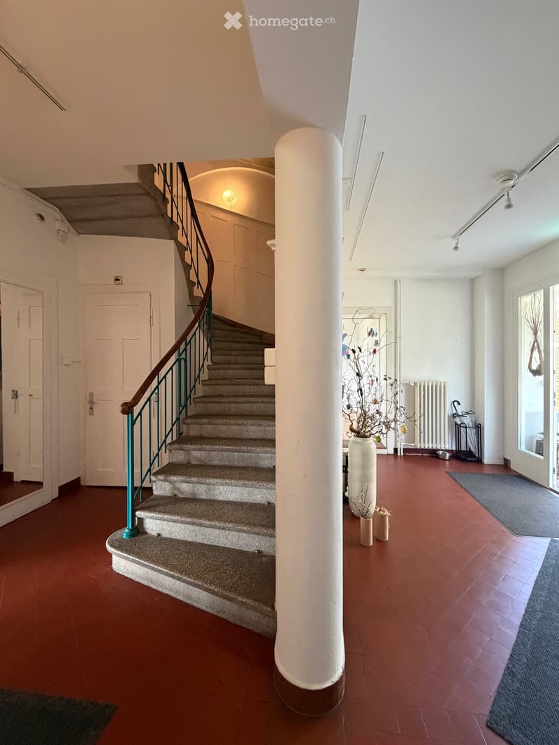 A hallway with a staircase, white walls, and red flooring. A white column stands in the center. A vase with branches is on the floor, and a radiator is against the wall. Rugs are placed on the floor, and an umbrella is near the door.
