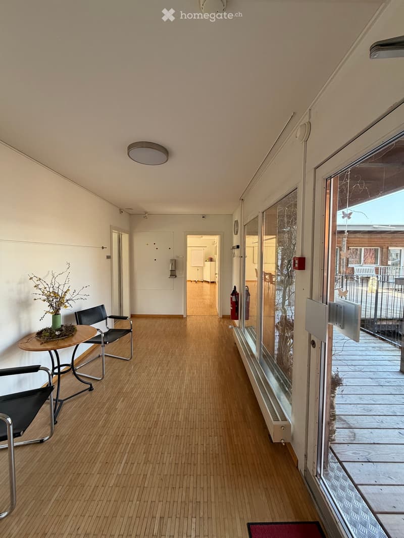 A corridor with wooden floors, white walls, and glass doors. There are two black chairs and a small table with a vase of flowers. A fire extinguisher is mounted on the wall, and a clock is visible. A radiator is installed on the wall, and a door is open.