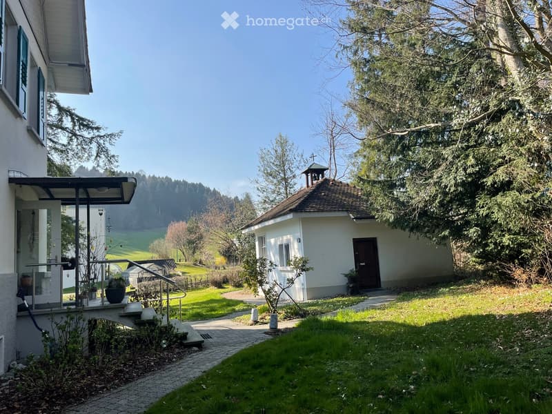 A small white house with a brown roof and a bell tower, surrounded by a well-maintained garden with a pathway leading to the entrance. The house is situated in a picturesque rural setting with lush greenery and a clear blue sky in the background.