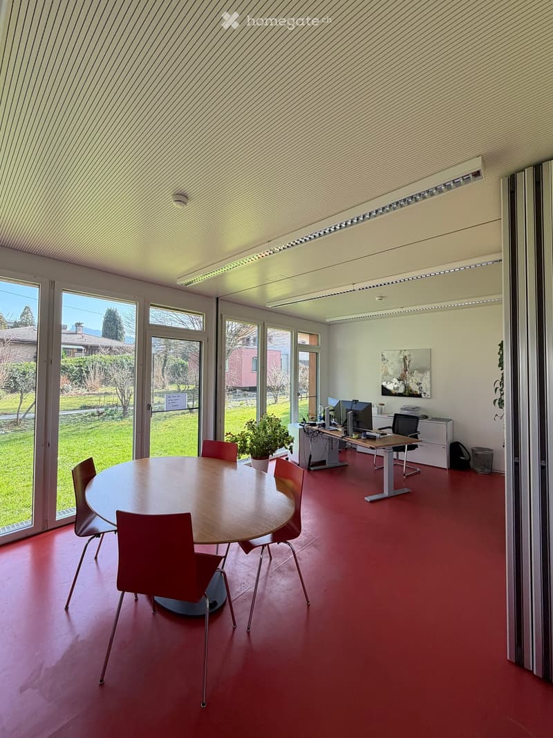 Modern office space with a round table, red chairs, and a desk with computers. Large windows provide natural light.