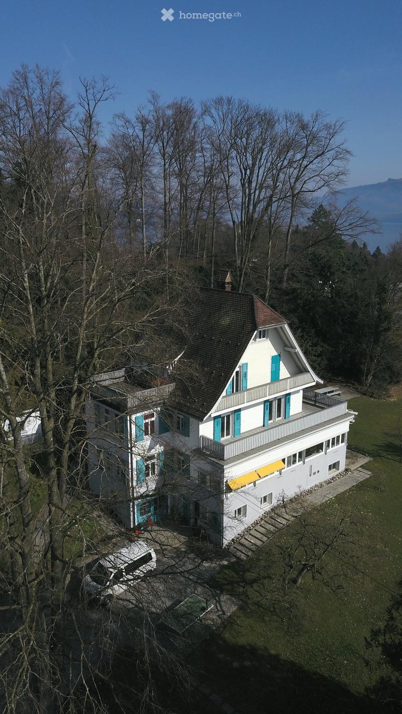 Aerial view of a large white house with a brown roof, blue shutters, and a yellow awning. The house is surrounded by trees and grass, with a car parked in front.
