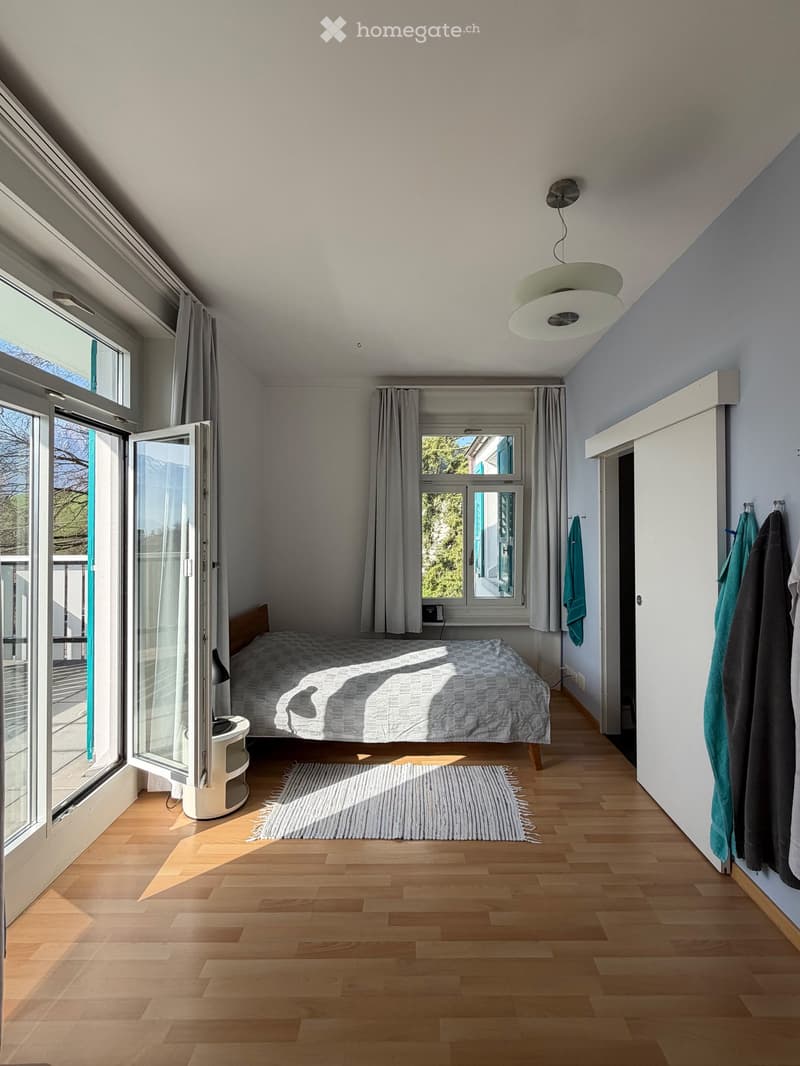 Spacious bedroom with wooden floor, large windows, and a bed. The room is furnished with a bed, a rug, and a lamp. There is a nightstand with a lamp on the left side of the bed. The room has a balcony with a view of the outside.