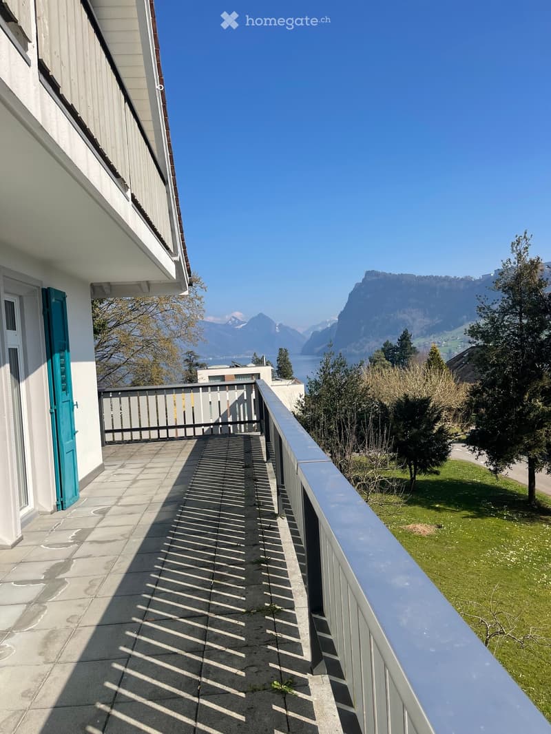 A balcony with a metal railing and a tiled floor, offering a view of a grassy area and mountains in the distance.