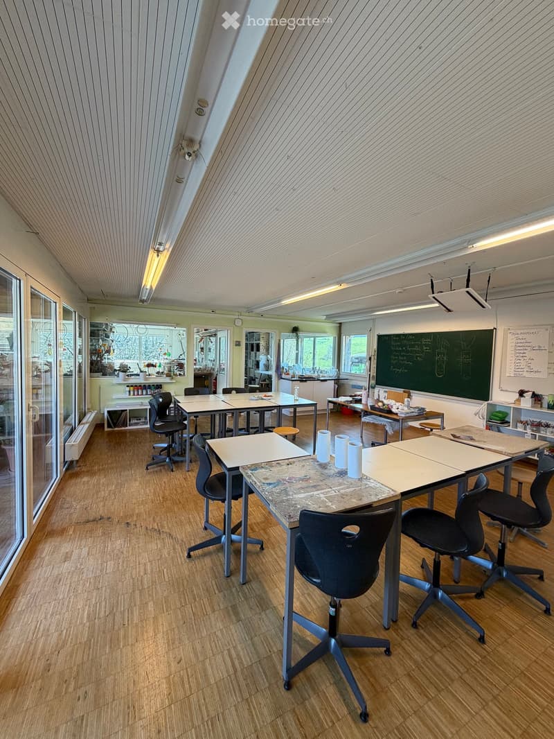 A classroom with desks, chairs, and a blackboard. The room has glass doors and windows, and a radiator is on the wall. There are shelves with various items, and a whiteboard is on the wall. The floor is wooden.