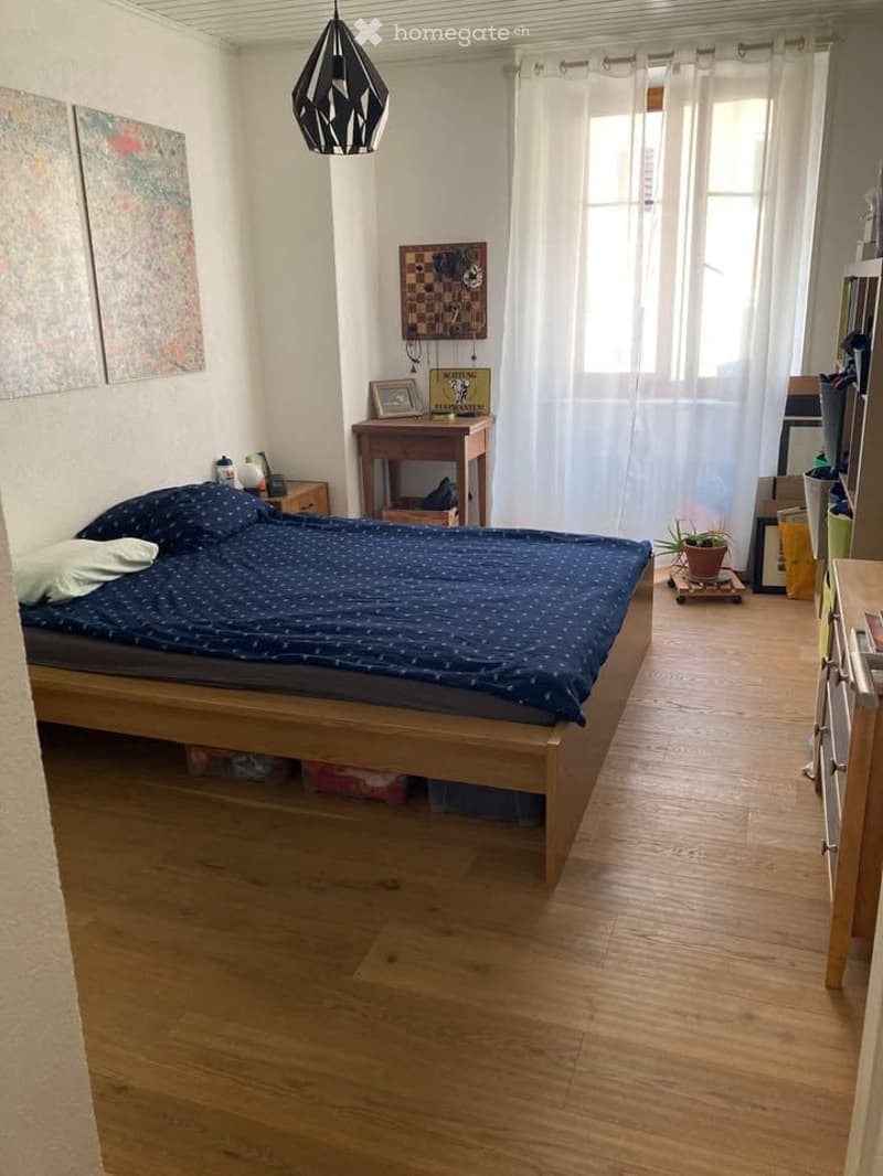 Cozy bedroom with wooden flooring, a double bed with blue bedding, a nightstand, a small table with a chair, a potted plant, and a pendant light fixture.