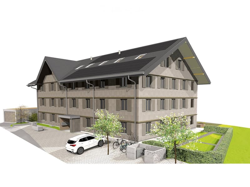 A modern multi-story residential building with a gray facade, multiple windows, and a sloped roof. The building features a covered parking area, bicycles, and landscaped greenery in front.
