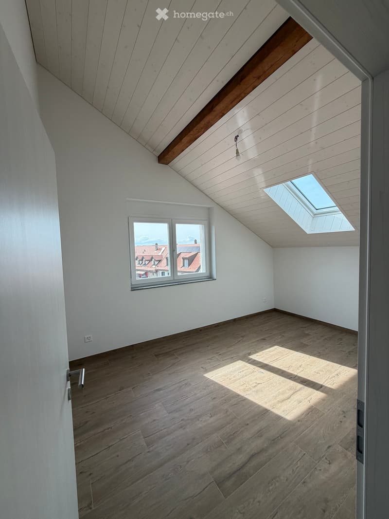 Empty attic room with wooden flooring, white walls, skylight window, and a regular window showing a view of rooftops.