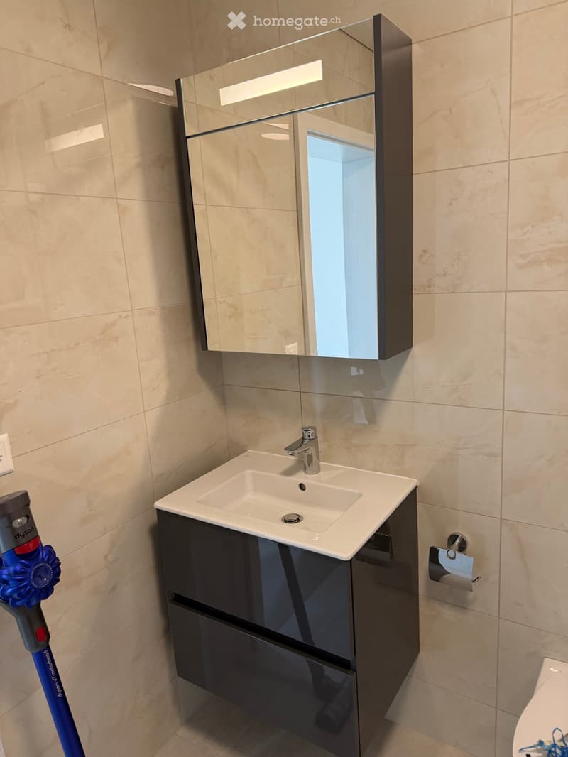 Modern bathroom with beige tiled walls, a white sink with a chrome faucet, a mirrored cabinet with built-in lighting, and a dark-colored cabinet beneath the sink.