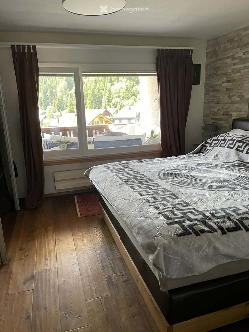 Cozy bedroom with wooden flooring, large window with view, bed with patterned duvet, and dark curtains.
