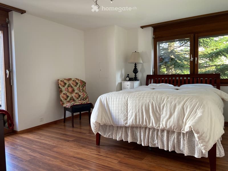 Cozy bedroom with wooden flooring, a large bed with white bedding, a patterned armchair, a nightstand with a lamp and clock, and large windows with a view of greenery.