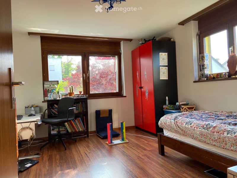A well-lit bedroom with a bed, desk, chair, bookshelf, and colorful decor items. The room features wooden flooring and large windows with a view of greenery.