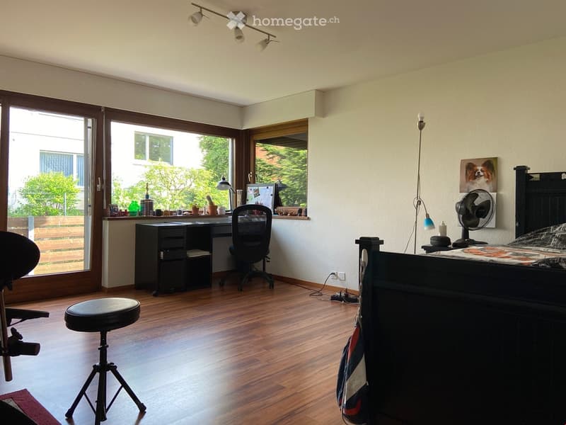 Well-lit room with wooden flooring, a bed, desk, chair, and a fan. Large windows and a door lead to an outdoor area.