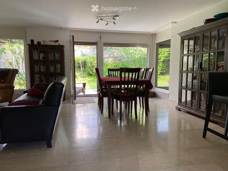 Open-plan living and dining area with a sofa, dining table, chairs, and cabinets. Large windows and sliding doors lead to a garden.