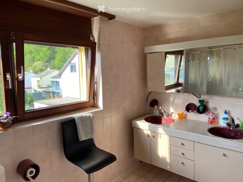 Bathroom with beige tiles, double sink, mirror, cabinet, and a window overlooking a garden. Includes a black chair, a flower pot, and a toilet paper holder.