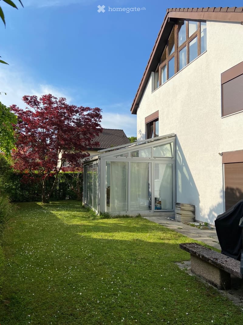 Well-maintained garden with a lawn, a stone bench, and a modern glass extension attached to a house. The house has a white facade with brown trim and multiple windows.