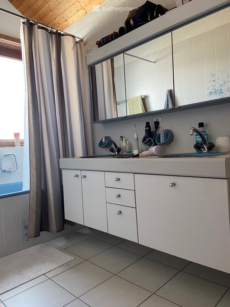 Modern bathroom with white tiled walls, a large sink with a mirror cabinet, and a shower area with a curtain. The sink has a faucet, toothbrush holder, and other toiletries. A hair dryer is placed on the counter, and a towel hangs on a rack.